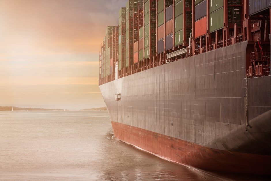 A colossal cargo ship loaded with containers navigates through calm waters against a vibrant sunset sky.